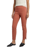 A model wearing slim-fit, ankle-length terracotta pants with side slits, paired with a cream ribbed top and black pointed mule flats, shown against a plain white background.