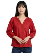 A woman wearing a red long-sleeve blouse with a split V-neckline, gathered shoulders, and button-front closure, paired with dark blue jeans against a plain white background.