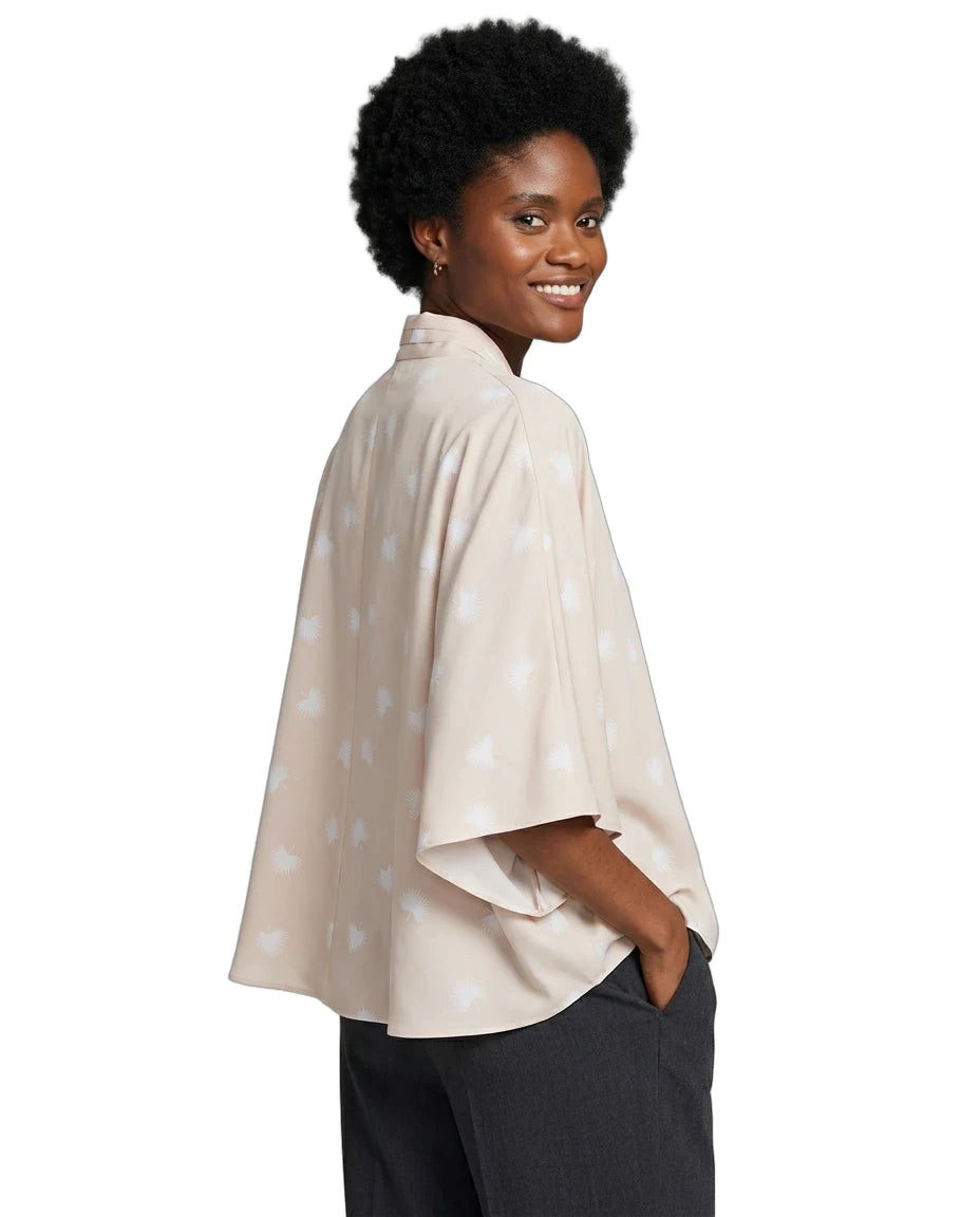 A woman with short black hair is shown from the back-three-quarter view wearing a light beige cape-style top with a subtle white star pattern, paired with dark gray pants, against a plain white background.