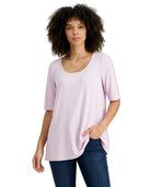 A woman with curly dark hair wearing a light lavender scoop-neck t-shirt and dark blue jeans, standing against a plain white background.