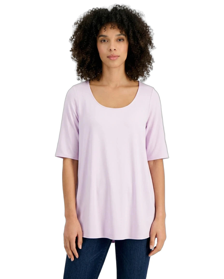 A woman with curly dark hair wearing a light lavender short-sleeve scoop-neck top and dark blue jeans, standing against a plain white background.