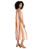 A woman modeling a sleeveless midi dress with vertical orange, peach, and pink watercolor-style stripes on a white base, shown from a side-back angle against a plain white background.