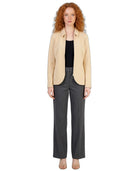A model with curly red hair stands against a white background wearing a light beige blazer over a black top, paired with dark gray wide-leg trousers and beige flat sandals.