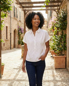 Woman in a white shirt and dark pants standing in a sunlit outdoor setting with plants and stone buildings.