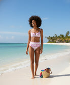Woman in a pink bikini standing on a beach with clear blue water and sky.