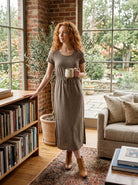 Woman in a brown dress standing in a cozy living room with a bookshelf and sofa.