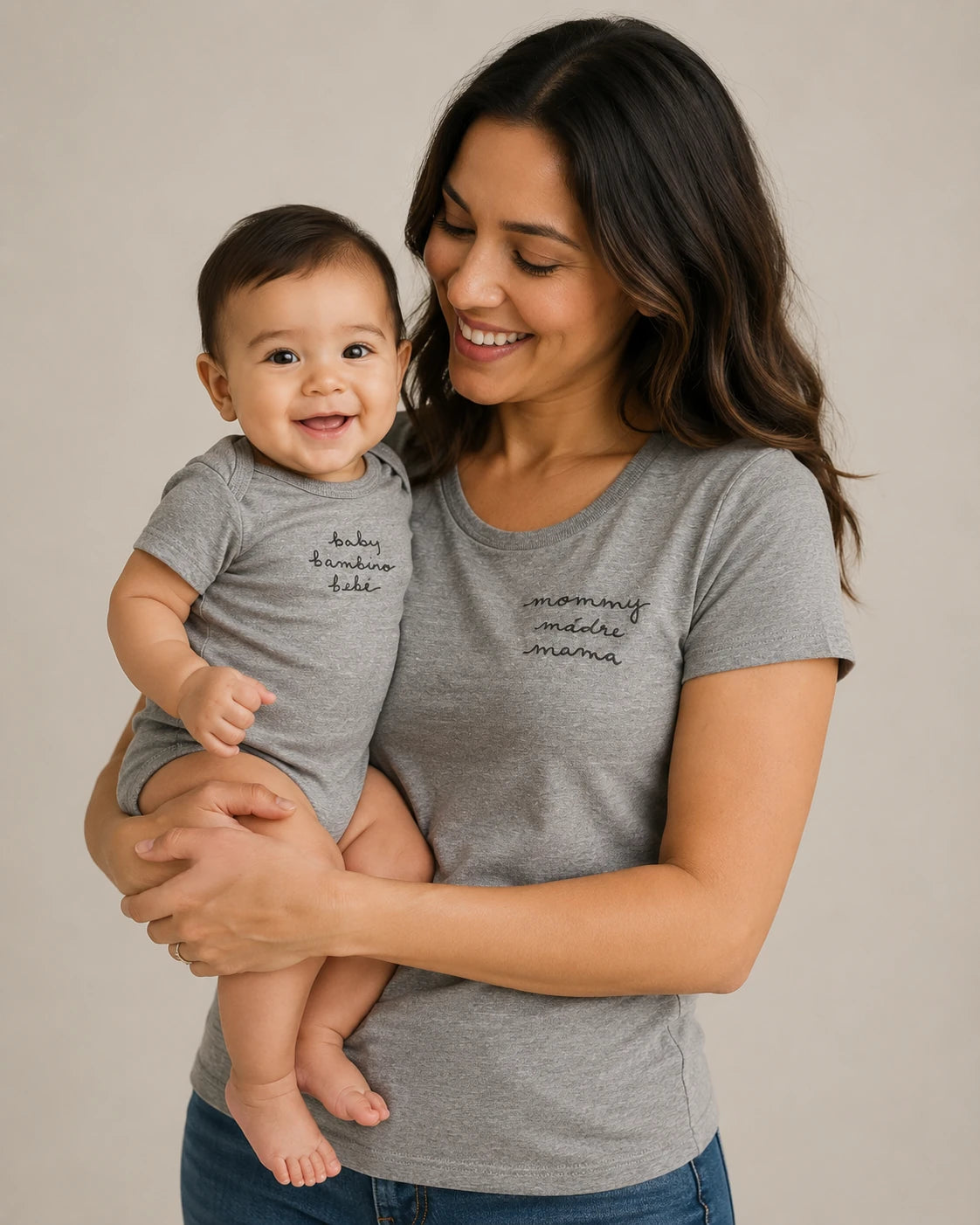 A woman holding a baby, both wearing matching heather gray t-shirts with black script text on the chest, photographed against a plain light beige studio background.